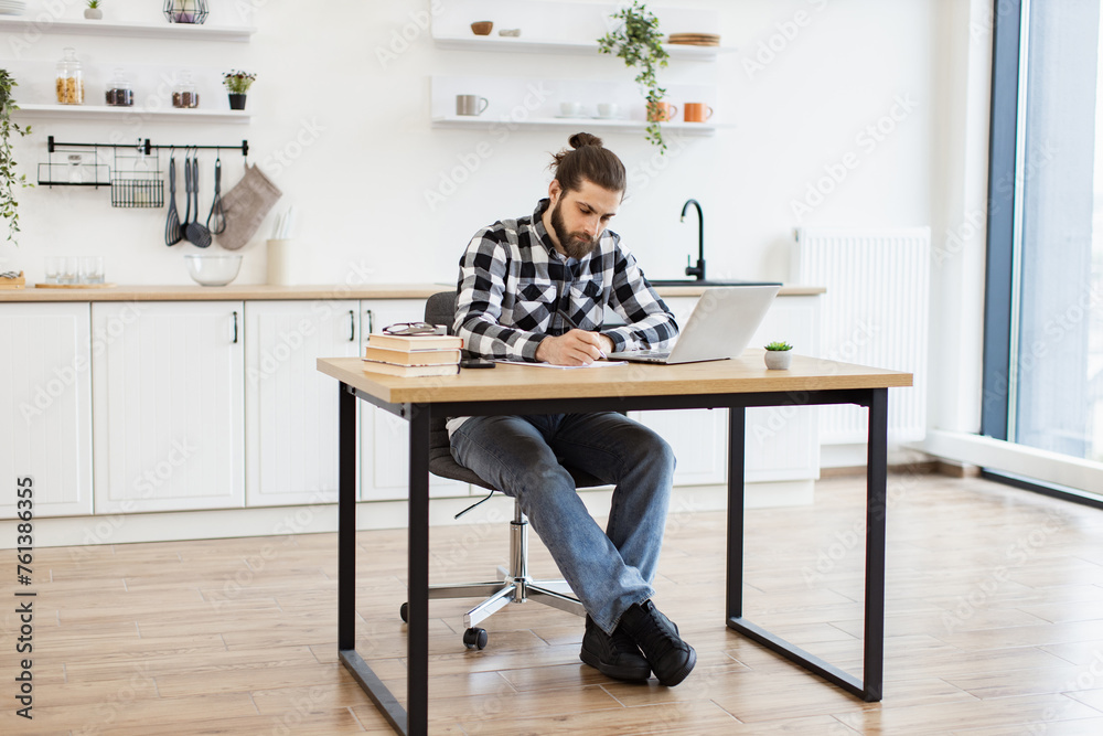 Full length of attractive Caucasian businessman sitting at office desk with digital devices in spacious dining room. Self-employed worker carrying out professional activity in comfortable workplace. Full length of attractive Caucasian businessman sitting at office desk with digital devices in spacious dining room. Self-employed worker carrying out professional activity in comfortable workplace.