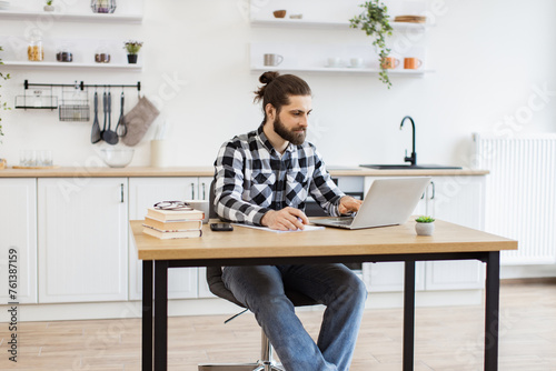 Bearded Caucasian businessman with pen making notes on draft of paper document being placed near laptop on wooden desk. Well-organised man remote employee starting business day in workplace at home.