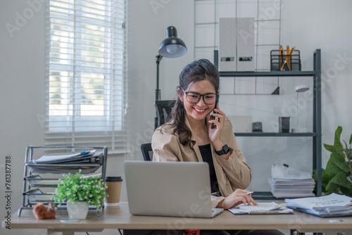 Young Asian business woman sits on the phone in an online business meeting using a laptop in a modern home office decorated with shady green plants.