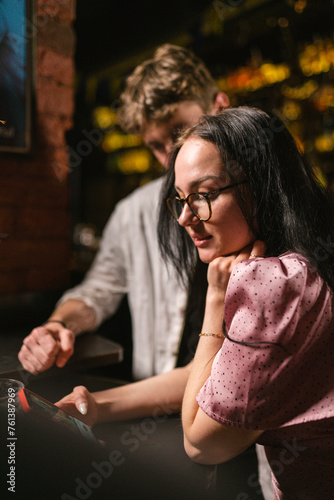 Woman in trendy dress wants to write secret message to new boyfriend at pub party. Guy sits near girl in glasses on blurred background