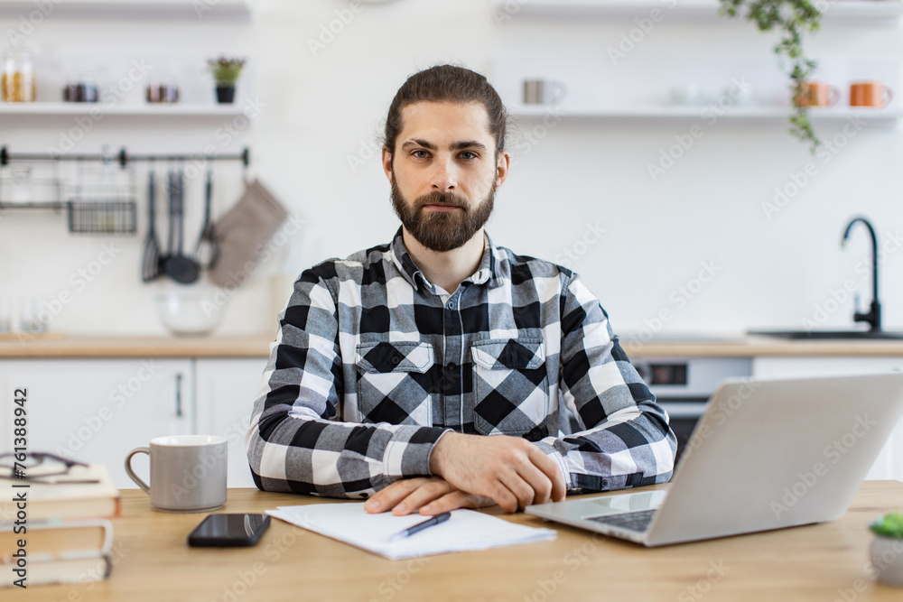 Confident Caucasian man in checkered shirt using laptop while sitting at desk on kitchen background, looking at camera. Efficient freelancer searching information while doing full-time job in home.