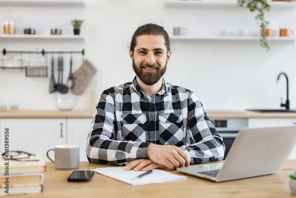 Fototapeta premium Confident Caucasian man in checkered shirt using laptop while sitting at desk on kitchen background, looking at camera. Efficient freelancer searching information while doing full-time job in home.