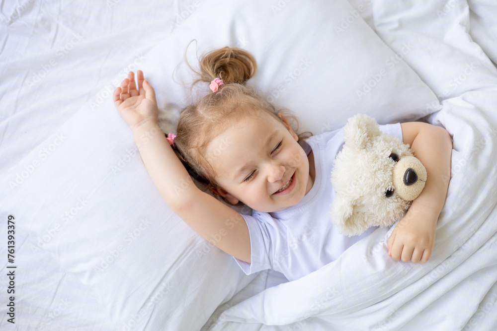 little cute blonde baby girl laughing in her sleep, sleeping on a white cotton bed at home hugging a teddy bear, close-up little cute blonde baby girl laughing in her sleep, sleeping on a white cotton bed at home hugging a teddy bear, close-up