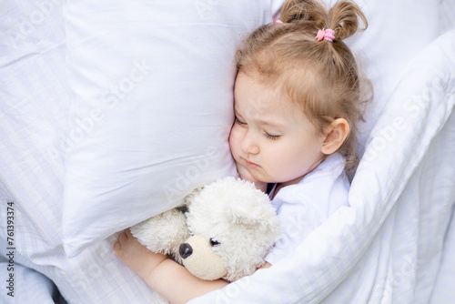 little cute blonde baby girl sleeping on a white cotton bed at home hugging a teddy bear, close-up