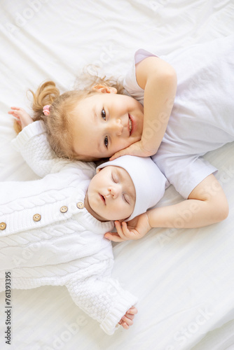 a little girl hugs a sleeping newborn sister or brother on a white bed at home, two children in the family