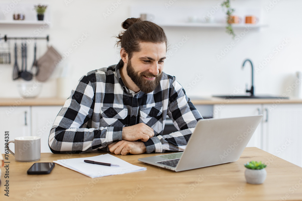 Smiling young man in casual clothes holding online conference with coworkers from home office. Positive Caucasian entrepreneur discussing company issues using portable computer in apartment.