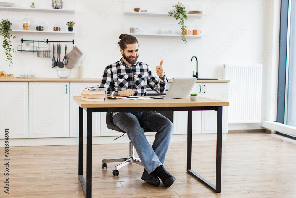 Positive Caucasian entrepreneur discussing company issues using portable computer in apartment. Smiling young man in casual clothes holding online conference with coworkers from home office. Positive Caucasian entrepreneur discussing company issues using portable computer in apartment. Smiling young man in casual clothes holding online conference with coworkers from home office.