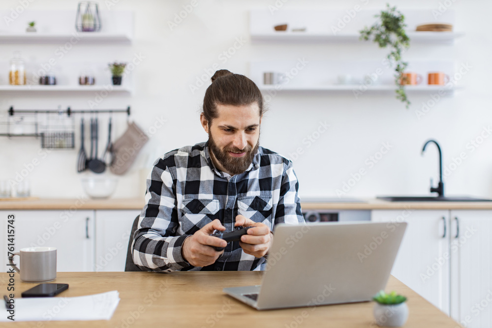 Concept of leisure, fun and entertainment at day time. Portrait of cheerful Caucasian guy in checkered shirt using remote console for playing video games at light kitchen at home. Concept of leisure, fun and entertainment at day time. Portrait of cheerful Caucasian guy in checkered shirt using remote console for playing video games at light kitchen at home.