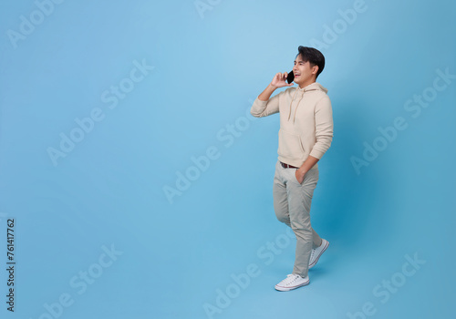 Young smiling happy asian man 20s talking speaking by mobile cell phone and walking isolated on blue background. portrait People lifestyle concept
