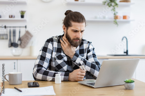 Sick young man in casual clothes touching sore throat while leveraging modern laptop in dining room. Deceased Caucasian employee working remotely from home while fighting illness indoors. © sofiko14