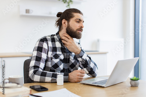 Deceased Caucasian employee working remotely from home while fighting illness indoors. Sick young man in casual clothes touching sore throat while leveraging modern laptop in dining room. photo