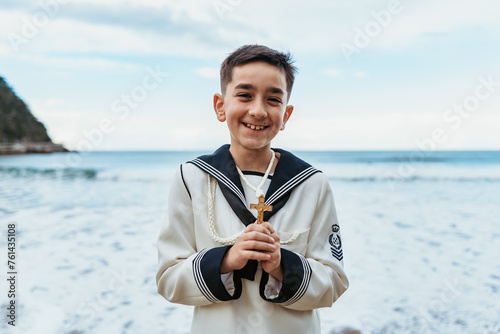 A beaming boy holding a crucifix stands by the sea, dressed in his First Communion sailor suit, embodying joy and faith photo