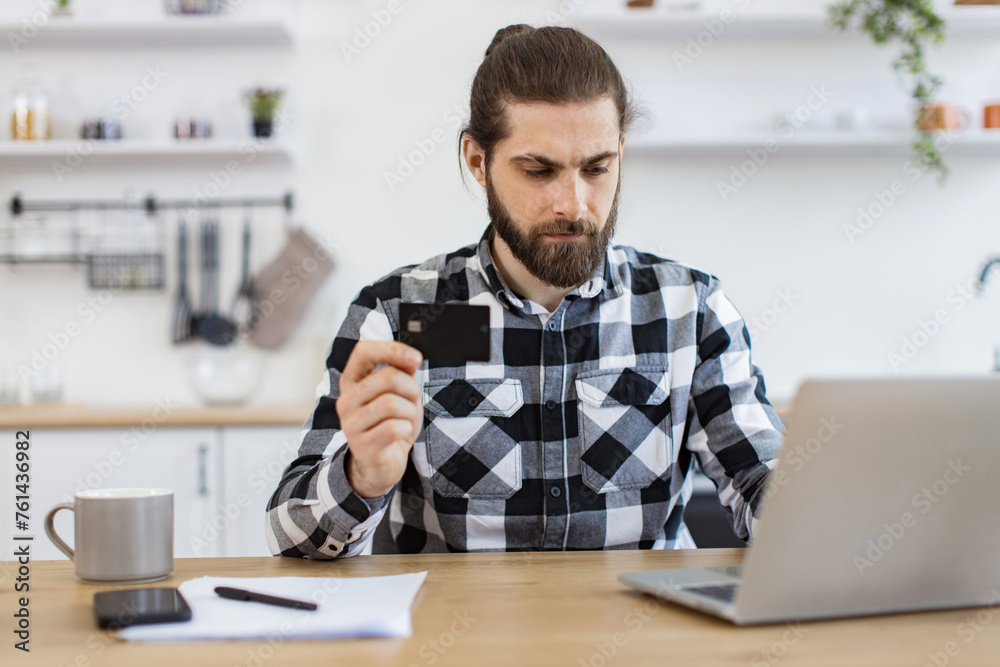 Attractive bearded man in checkered shirt holding credit card while typing on portable computer in kitchen interior. Happy Caucasian gentleman doing online banking transaction while working remotely.