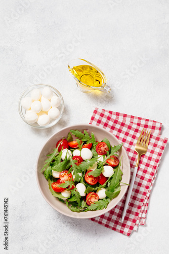 Fresh green salad made with mozzarella cheese, cherry tomatoes, arugula and olive oil on white stone table background. Healthy food and diet concept. Flat lay, top view, copy space.