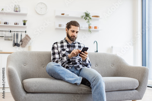 Caucasian male in checkered shirt holding smartphone while staying in modern workplace at home. Efficient freelance worker texting message to colleague while performing project for employer indoors.