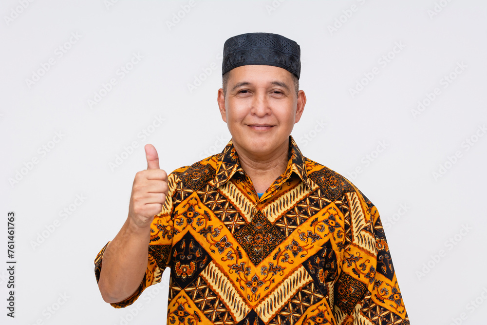 Portrait of a cheerful Indonesian man wearing a traditional batik shirt and songkok, giving a thumbs-up gesture on a white background. Portrait of a cheerful Indonesian man wearing a traditional batik shirt and songkok, giving a thumbs-up gesture on a white background.