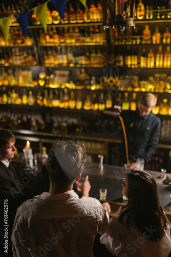 Young customers wait for alcoholic cocktails sitting at bar counter. Concentrated bartender in work uniform makes drinks with aluminum shaker pouring orange liquid