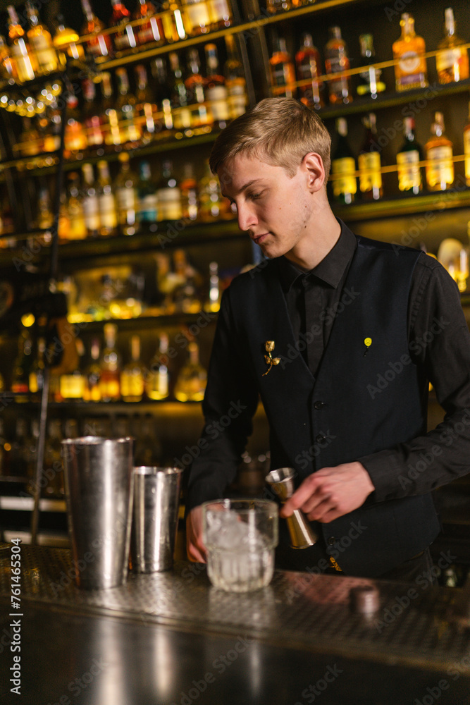 Blonde bartender prepares workplace for working shift in bar. Young barman with jigger in hand looks for bottle of alcohol for first order of evening