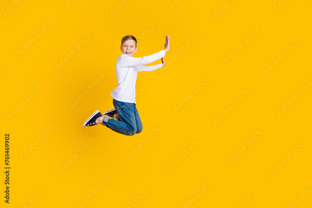 Full length photo of excited funky kid dressed white shirt jumping high pushing arms hands empty space isolated yellow color background