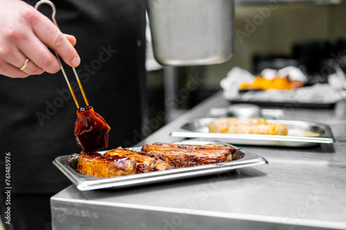 Professional chef in a kitchen glazing freshly grilled ribs with a thick sauce, with other food trays in the background.