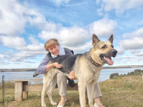 An adult girl with a shepherd dog on a bench by a river or lake. Middle aged woman and big shepherd dog on nature. Friendship, love, communication, fun, hugs © keleny