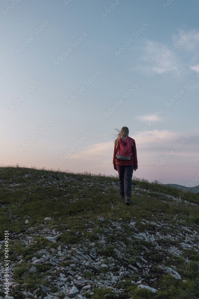 Female hiker silhouette with a backpack walking and following the trail uphill, enjoying the sky at sunset. Inspiration and healthy lifestyles concepts.