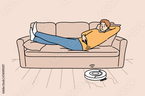 Robot vacuum cleaner is controlled using remote control, cleaning room with guy lying on sofa. Modern vacuum cleaner with wifi connectivity that independently cleans floor in apartment