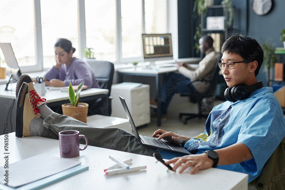 Side view portrait of Asian young man working with laptop while relaxing casually at workplace in office copy space Side view portrait of Asian young man working with laptop while relaxing casually at workplace in office copy space