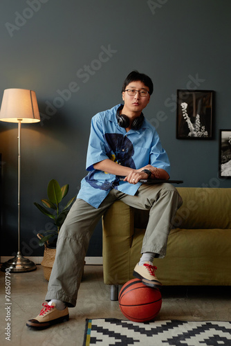 Vertical full length portrait of Asian young man working from home or in office lounge and looking at camera playing with basketball