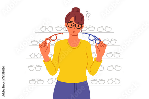 Woman buys glasses in store with large assortment of lenses and frames and chooses one of two options. Girl stands near display case with glasses in ophthalmic shop, allowing to solve vision problems.