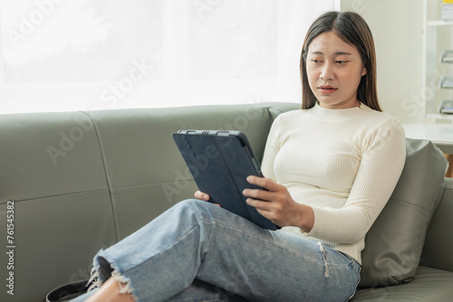 Attractive young Asian woman sitting on gray sofa and using digital tablet at living room, texting message on tablet. Smiling young woman uses a tablet online message chat Shopping online from home