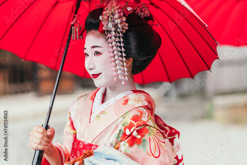 A geisha under a vermilion umbrella presents a calm demeanor, her pink kimono adorned with intricate flowers. Her look captures the essence of Japanese artistic tradition photo