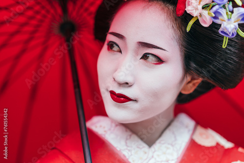 A geisha's contemplative gaze peeks from beneath her red umbrella, surrounded by the textures of her traditional attire. The image evokes a sense of quiet introspection. photo