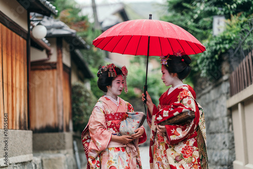 A candid shot of two Geishas sharing a lighthearted conversation, the red umbrella above them symbolizing their shared tradition photo