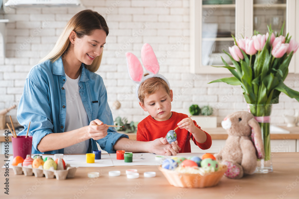 Happy positive mother and little son decorating eggs for Easter at home. Mom and her small kid child painting ornaments for holiday celebration together in the kitchen