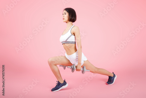 Sporty woman holding two dumbells and doing leg lunges for leg muscle training isolated on pink background. Woman with a healthy lifestyle.