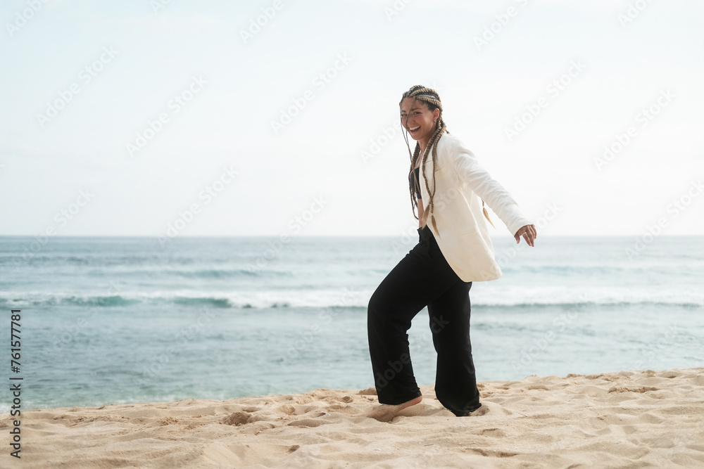 Medium shot of a young woman with braids wearing black trousers and a white jacket walking along the beach near the ocean and laughing. The concept of masculine clothing style. Medium shot of a young woman with braids wearing black trousers and a white jacket walking along the beach near the ocean and laughing. The concept of masculine clothing style.