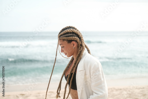 Portrait of a young Latino woman with braids on her head. She walks along the beach near the ocean in a jacket and suit. Self confidence and masculinity concept. Braiding studio.