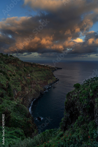 A serene view of a rocky coast with red flowers, under a dusk sky with fluffy clouds Colorful sunset at Caniço, Madeira island, Portugal