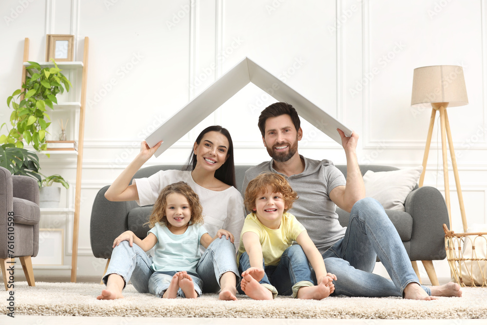 Family housing concept. Happy woman and her husband holding plastic roof while sitting with kids on floor at home Family housing concept. Happy woman and her husband holding plastic roof while sitting with kids on floor at home