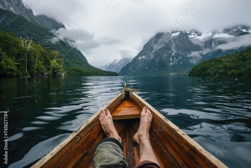 A person's bare feet propped up in the foreground, relaxing on the edge of a wooden boat, with a serene lake and mountains in the distance