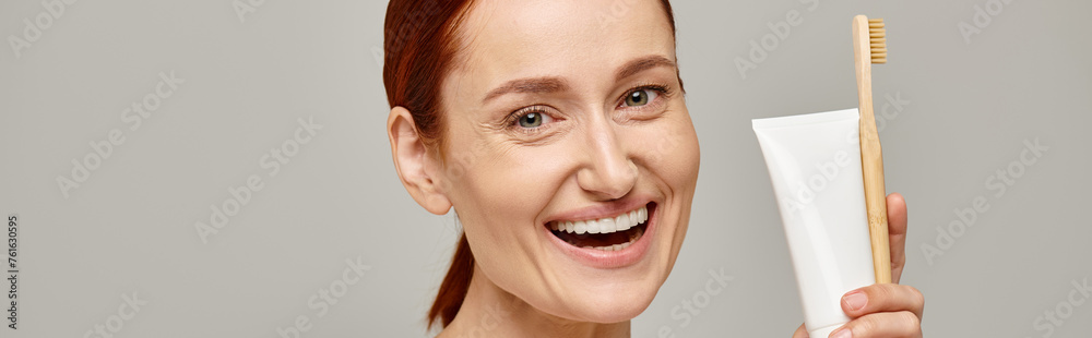 excited woman with red hair holding toothpaste and toothbrush and smiling at camera on grey, banner excited woman with red hair holding toothpaste and toothbrush and smiling at camera on grey, banner