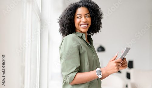 Beaming with happiness, an African-American woman holds and operates a tablet, her engagement with the device suggesting a pleasant online experience in a comfortable and bright interior setting