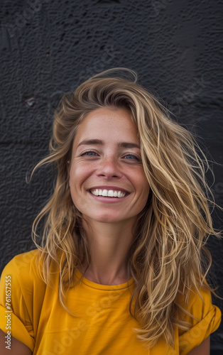A beautiful happy and smiling young blonde woman in yellow T-shirt, behind her a black concrete wall © Giordano Aita
