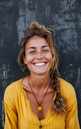 A beautiful happy and smiling young blonde woman in yellow shirt, behind her a black concrete wall © Giordano Aita