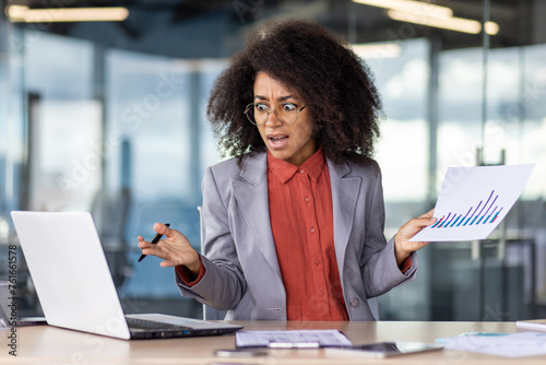 Photos Confused head of department holding sheet with charts and pen in raised hands while looking at pc screen with open mouth