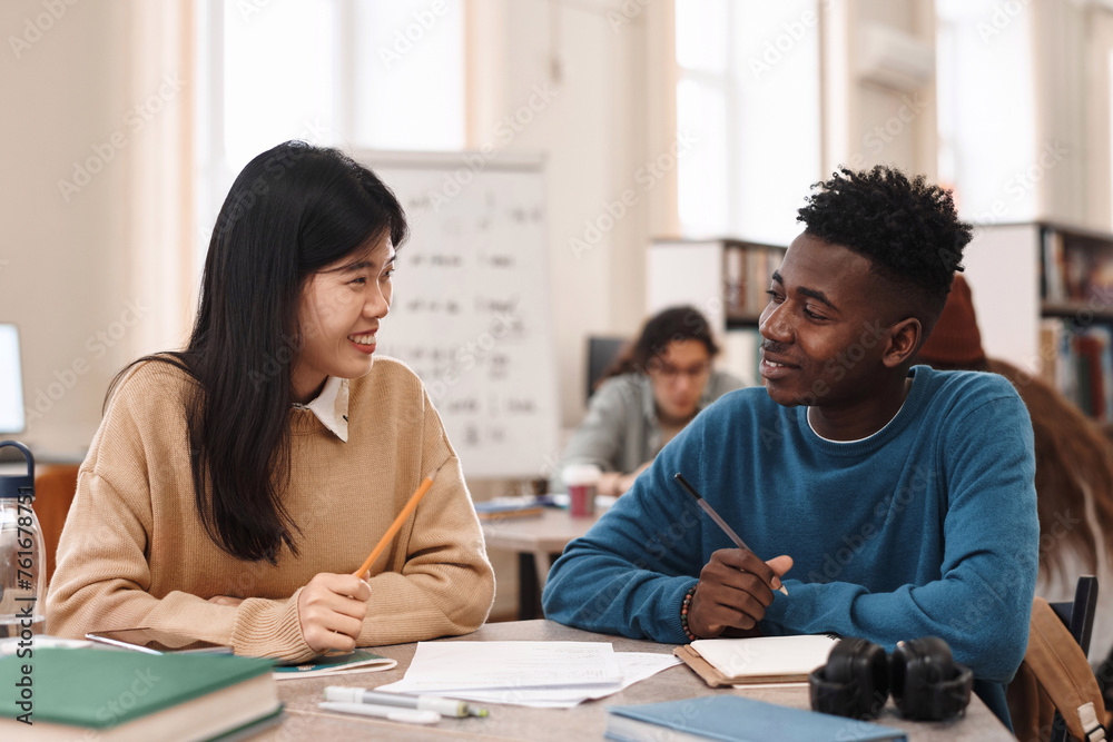 Side view portrait of smiling Asian woman looking at Black student while studying together in college library Side view portrait of smiling Asian woman looking at Black student while studying together in college library
