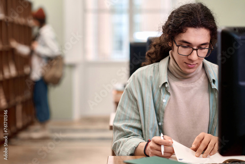 Front view portrait of young male student wearing glasses writing in notebook while studying in college library copy space