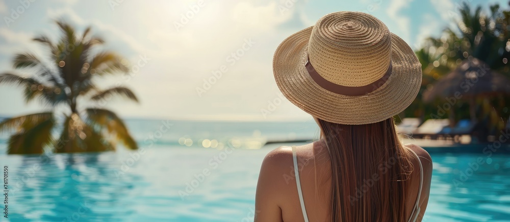 A woman sporting a sun hat is relaxing by a pool gazing at the liquid expanse of the ocean beneath the vast sky, enjoying leisure in a picturesque landscape