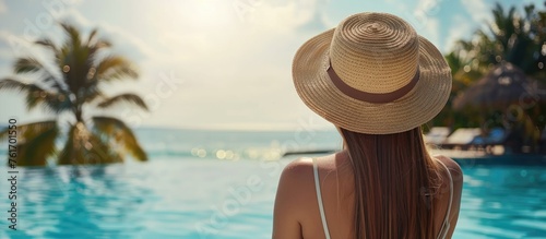 A woman sporting a sun hat is relaxing by a pool gazing at the liquid expanse of the ocean beneath the vast sky, enjoying leisure in a picturesque landscape © 2rogan